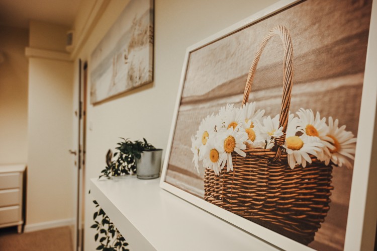 Zdjęcie przedstawia widok z kanapy na ścianę na której zawieszony jest obraz rumianków w wiklinowym koszu. 